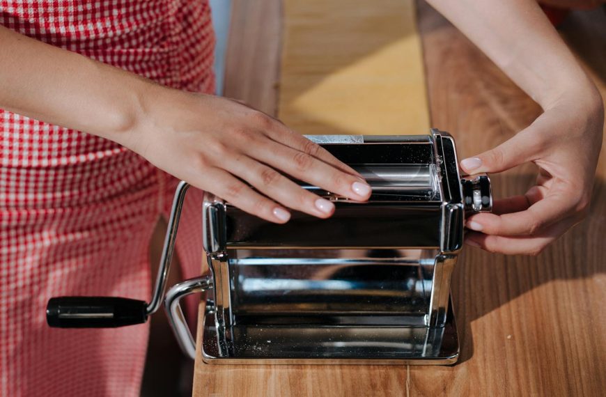 Hands making homemade pasta using a shiny metal pasta maker on a wooden table.