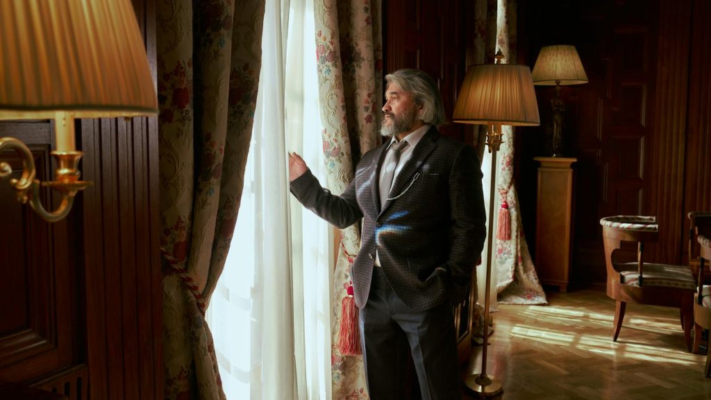 Senior man in a suit stands by window in elegantly decorated luxury interior of a mansion.