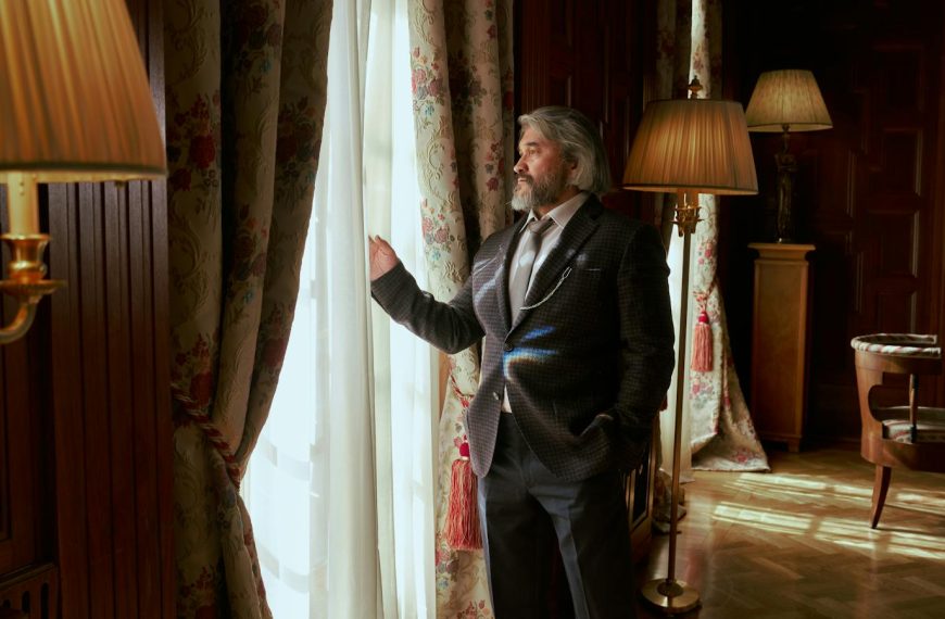 Senior man in a suit stands by window in elegantly decorated luxury interior of a mansion.