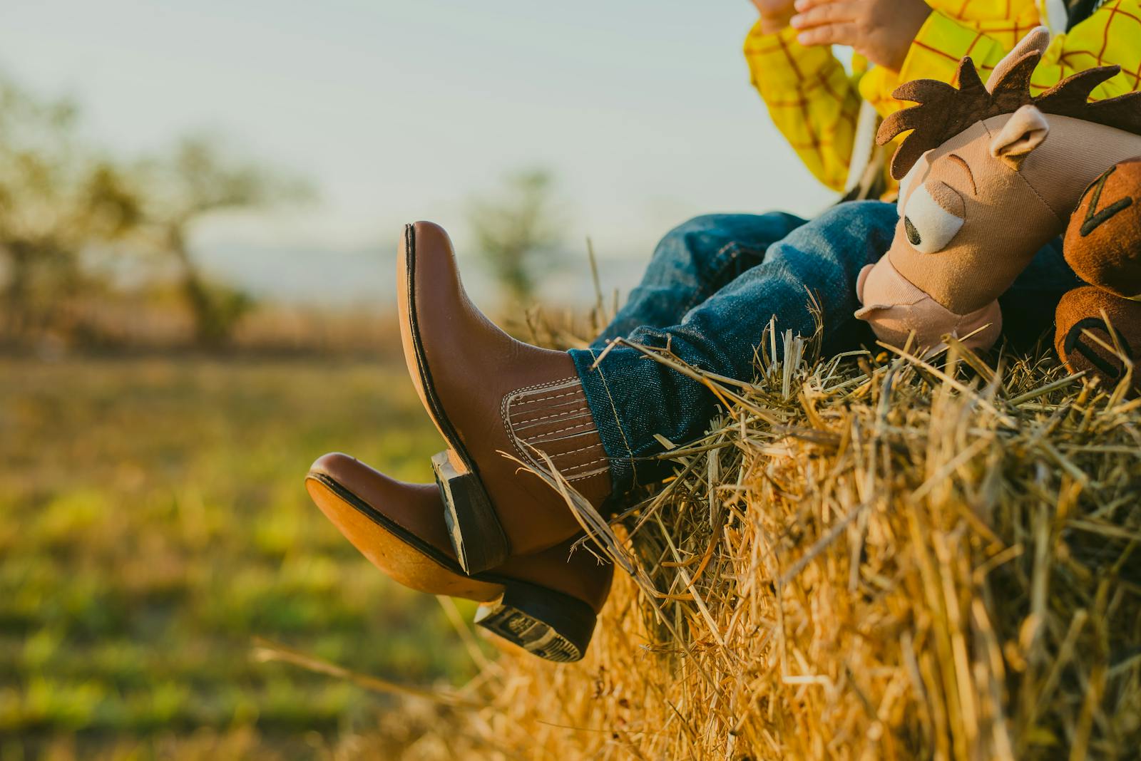 A child in jeans and boots sitting on a haystack with a toy, in a sunlit field.