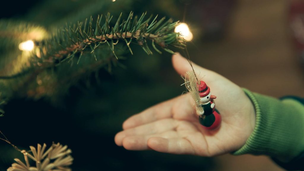 A close-up of a child placing a snowman ornament on a Christmas tree branch with lights.