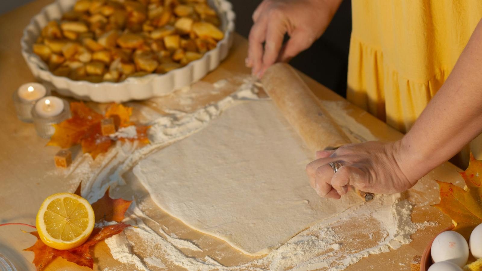 Woman rolling dough for apple pie surrounded by autumn decor and ingredients.