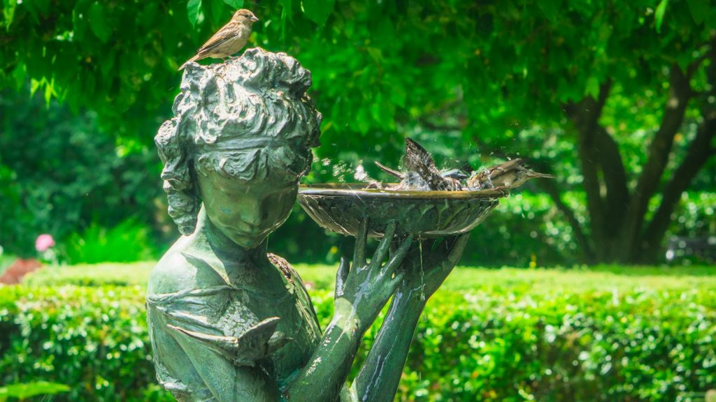 A serene bronze statue in a lush New York garden with birds enjoying a bath under sunlight.