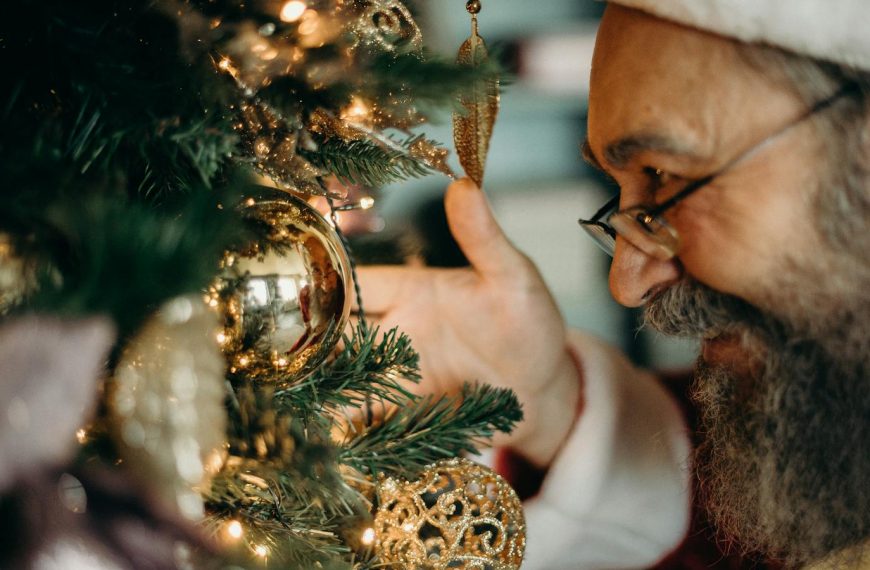 Elderly man admiring Christmas tree ornaments with a joyful expression.