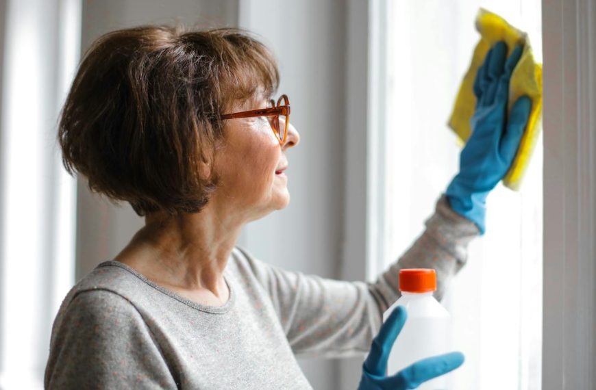 Elderly woman cleansing window indoors using gloves and cleaner.