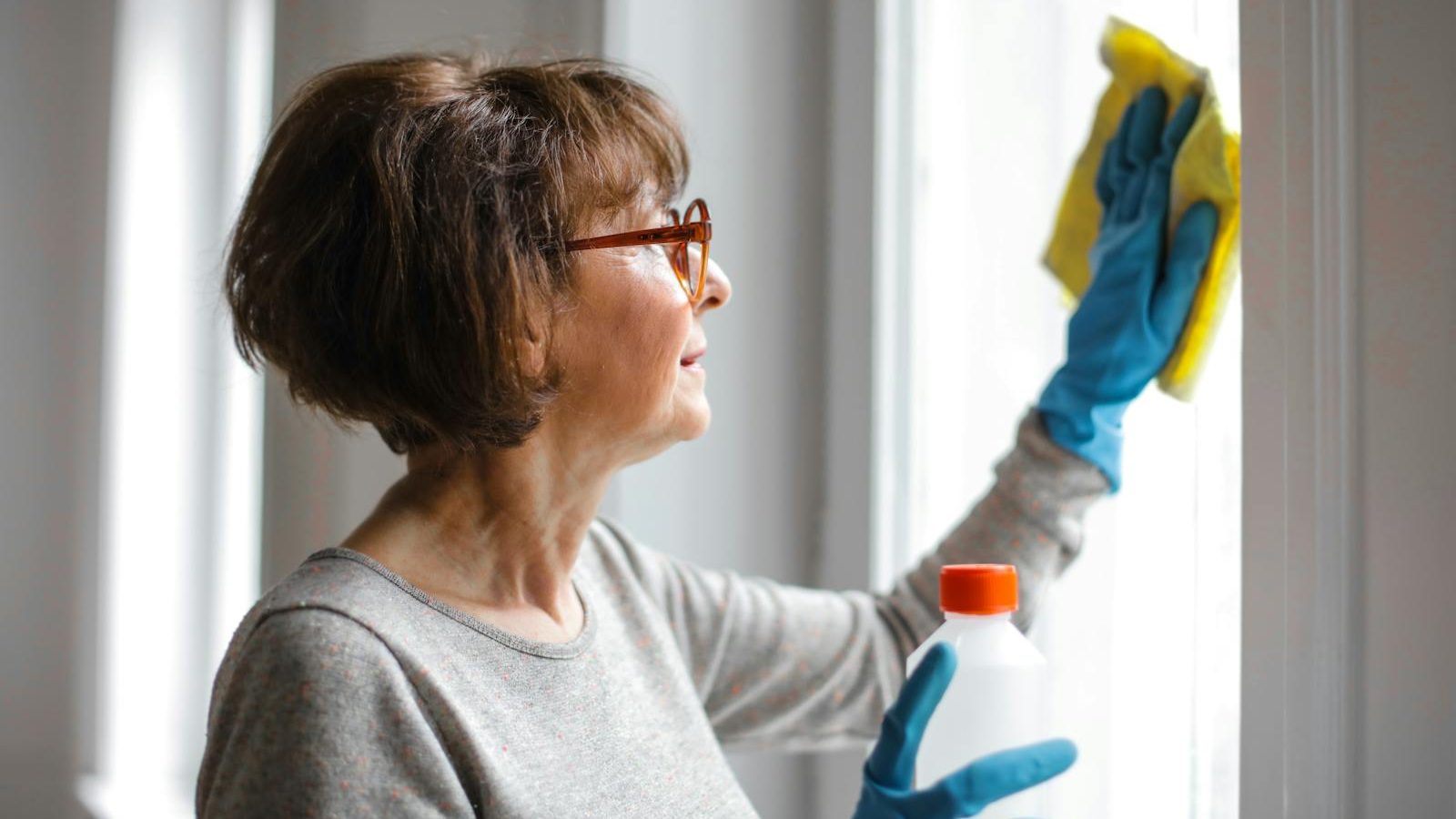 Elderly woman cleansing window indoors using gloves and cleaner.