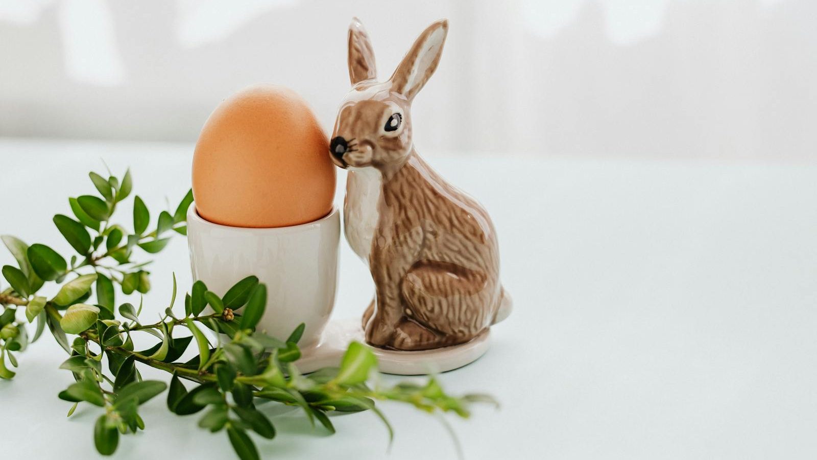 A festive Easter scene with a ceramic bunny, egg in holder, and greenery on a light background.