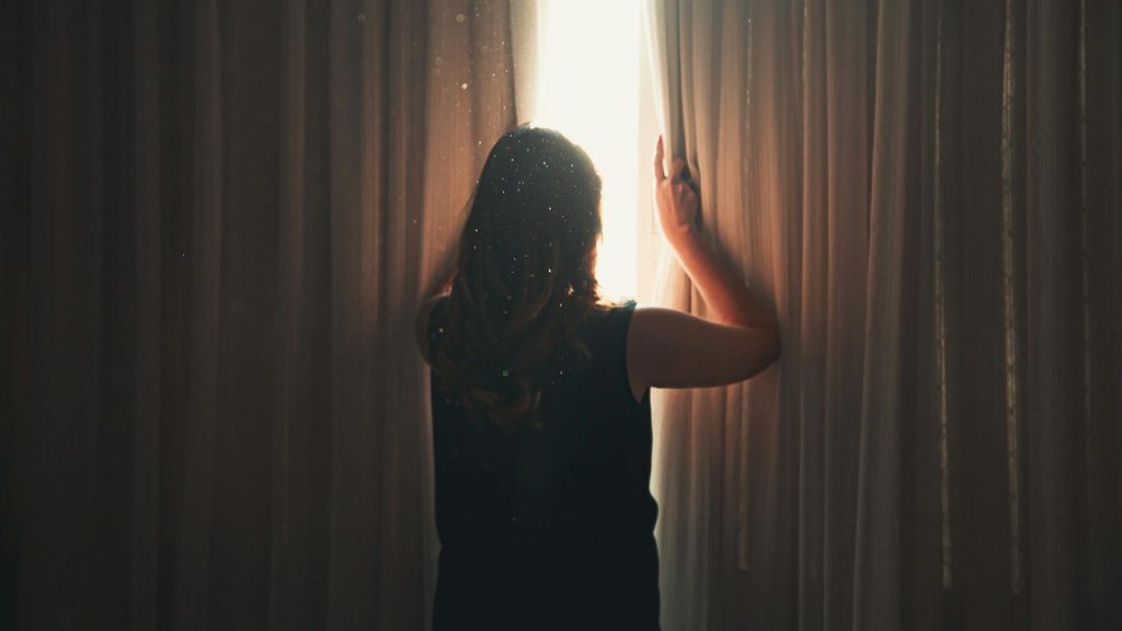 Back view of anonymous lady opening ribbed curtain behind window with glowing light while standing in apartment