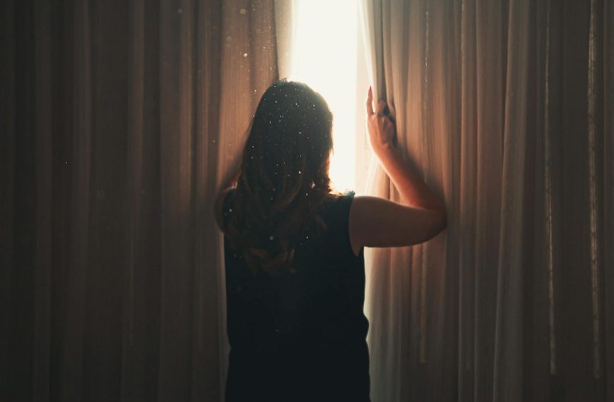 Back view of anonymous lady opening ribbed curtain behind window with glowing light while standing in apartment