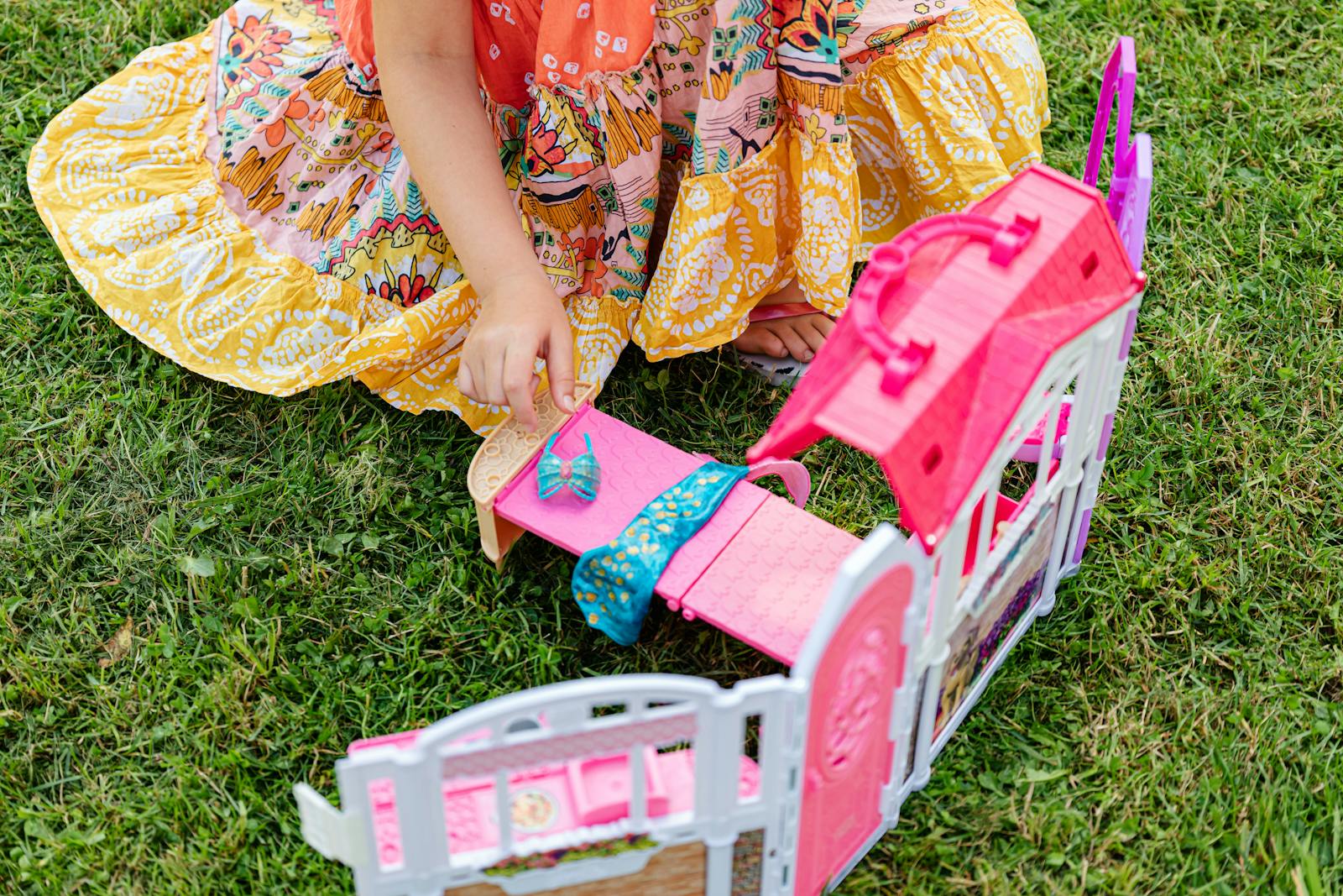 A child playing outside with a colorful floral dress and a toy house on grass.