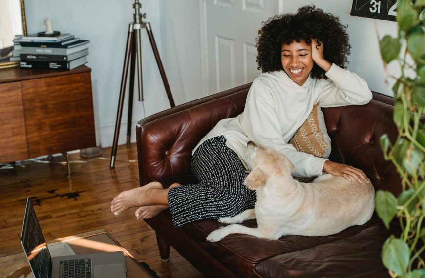 Happy young African American woman sitting on couch and resting with adorable dog with computer and cup of coffee on table in living room