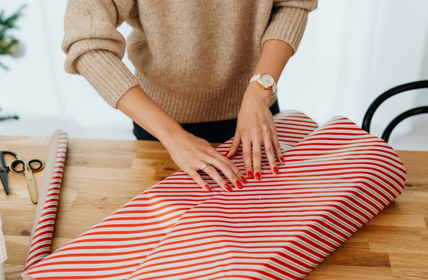 A woman wrapping a Christmas gift indoors using red and white striped paper, surrounded by holiday decorations.