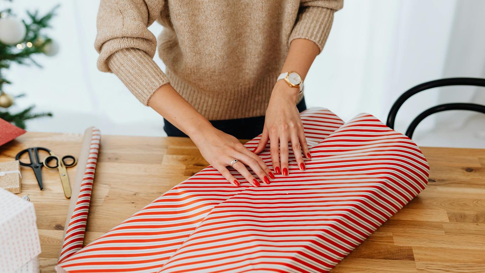 A woman wrapping a Christmas gift indoors using red and white striped paper, surrounded by holiday decorations.