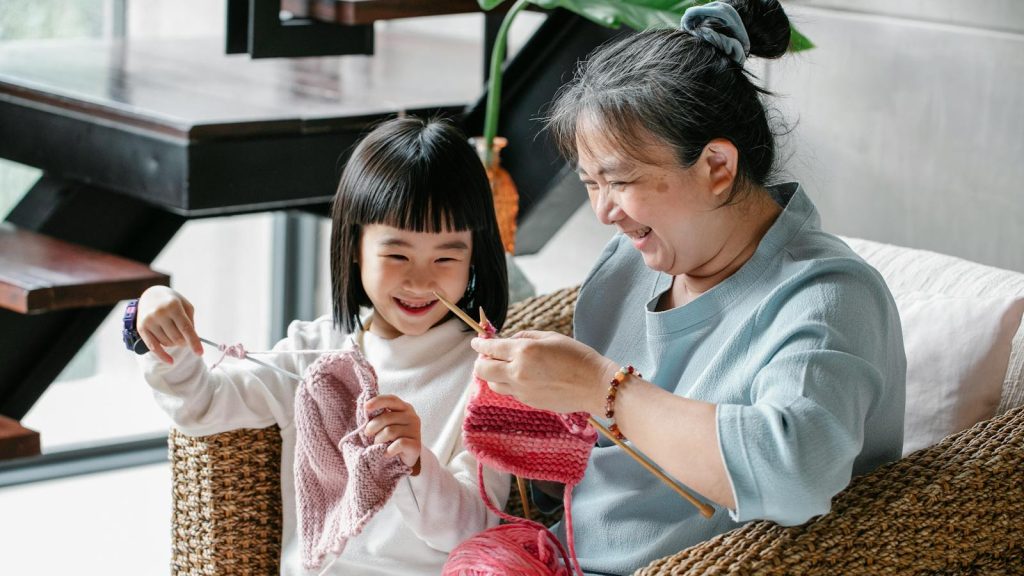 Asian grandmother teaching young granddaughter to knit in cozy home setting.