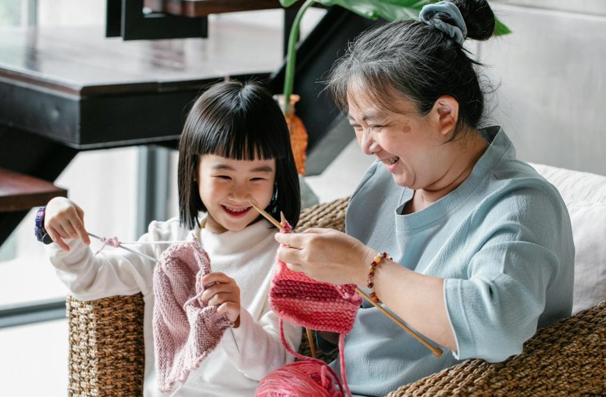 Asian grandmother teaching young granddaughter to knit in cozy home setting.