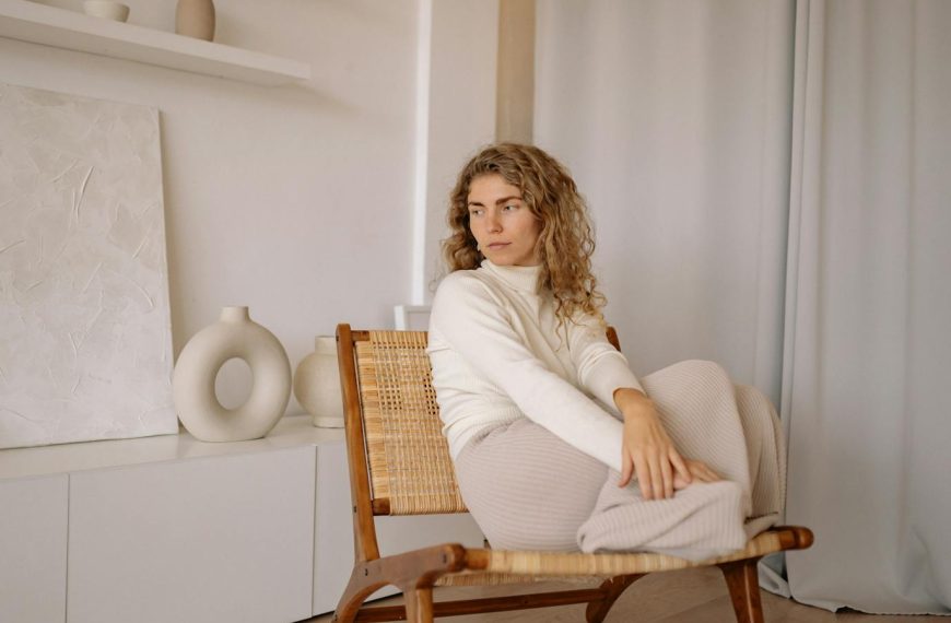 A woman in a minimalist room, sitting on a wicker chair by the curtain, surrounded by vases and art.