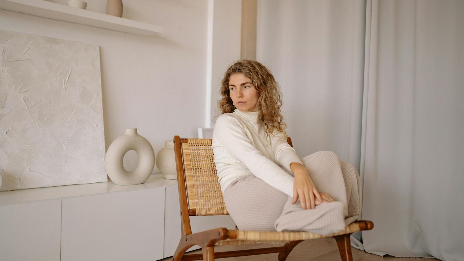 A woman in a minimalist room, sitting on a wicker chair by the curtain, surrounded by vases and art.