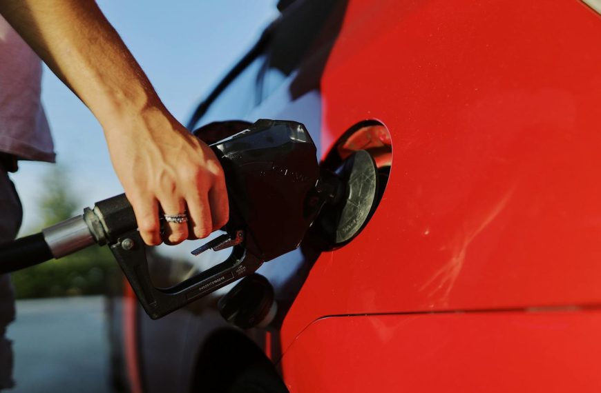Close-up of a person refueling a red car at an outdoor gas station during the day.