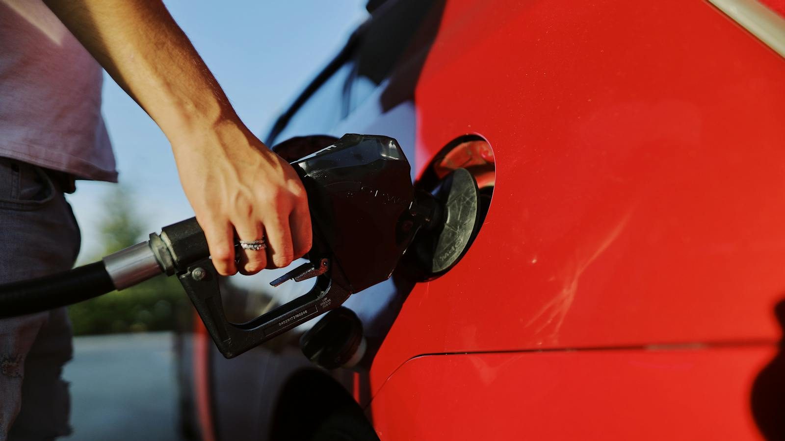 Close-up of a person refueling a red car at an outdoor gas station during the day.