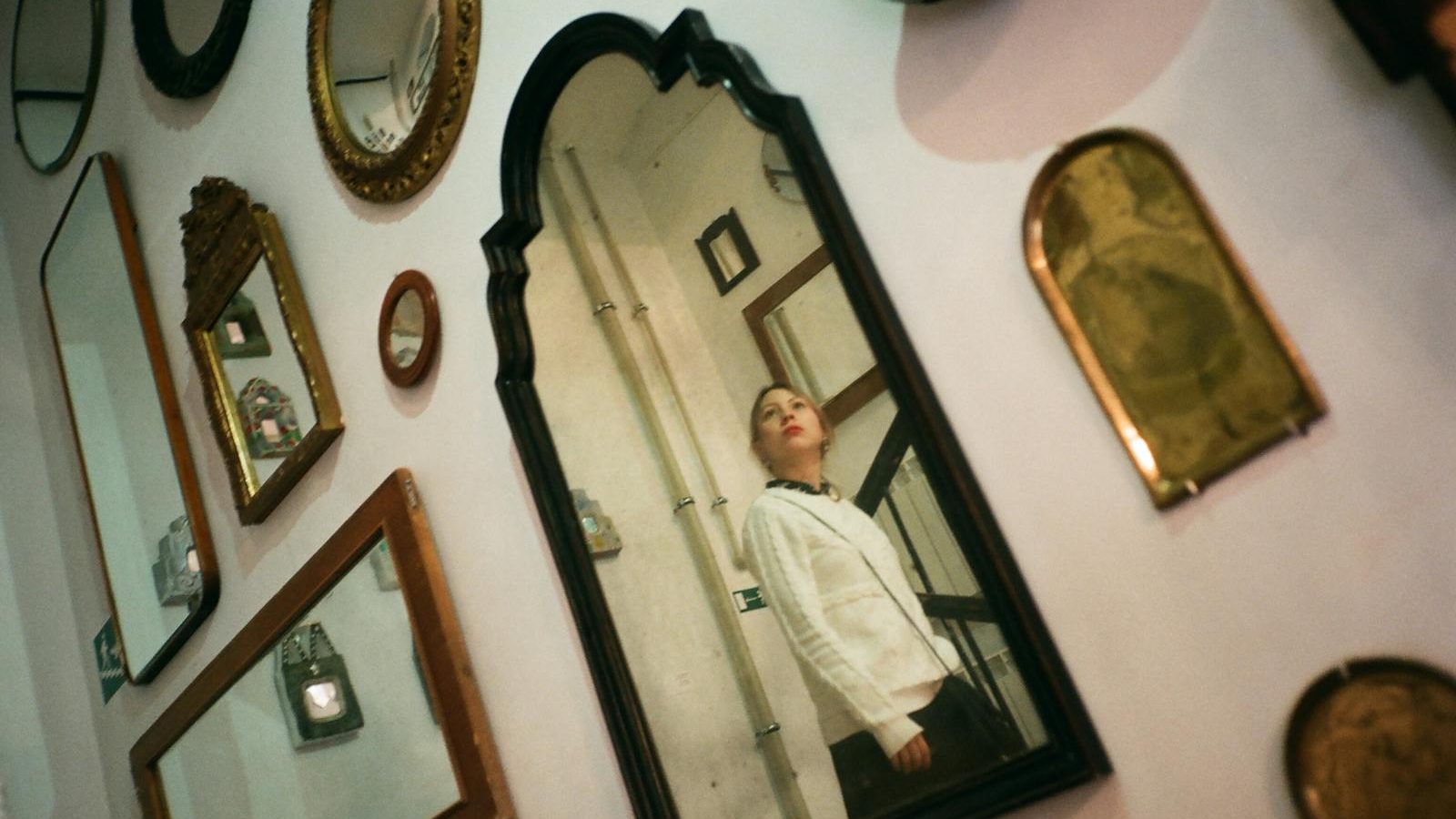 Contemplative young female in casual wear standing on stairs and reflecting in one of many framed mirrors hanging on wall
