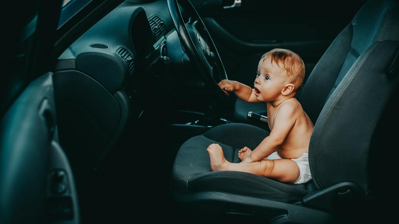 Cute baby sitting on the driver's seat of a car, looking curious and playful.