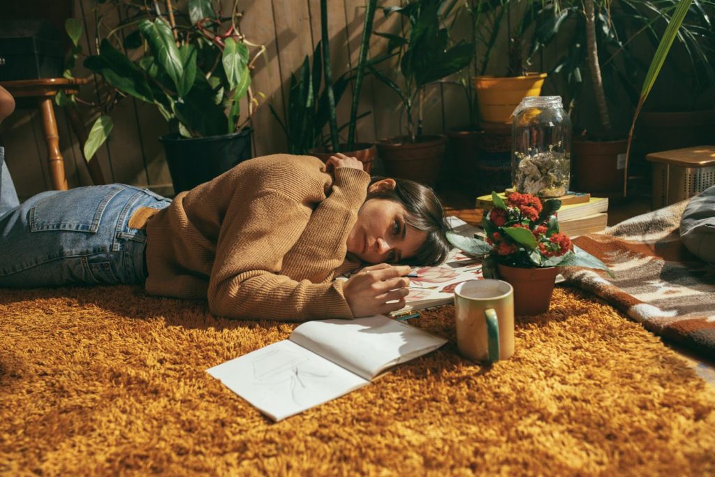 Woman in sweater lying on carpet drawing, surrounded by plants and books, enjoying leisure time indoors.