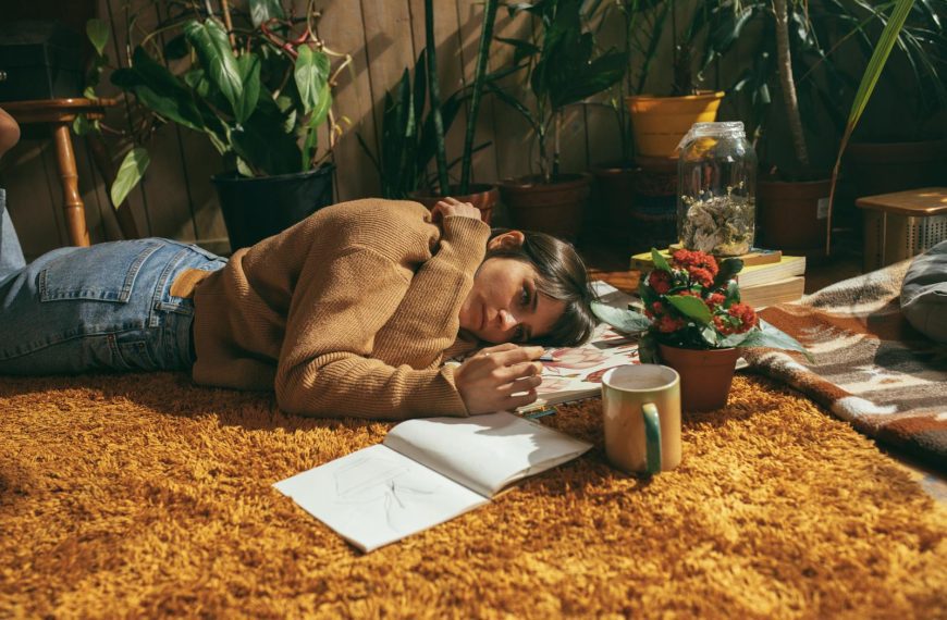 Woman in sweater lying on carpet drawing, surrounded by plants and books, enjoying leisure time indoors.