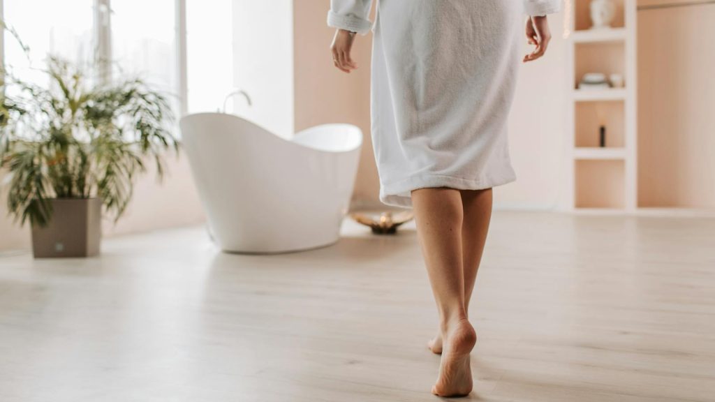A person in a bathrobe walks towards a stylish bathtub in a bright, modern bathroom.