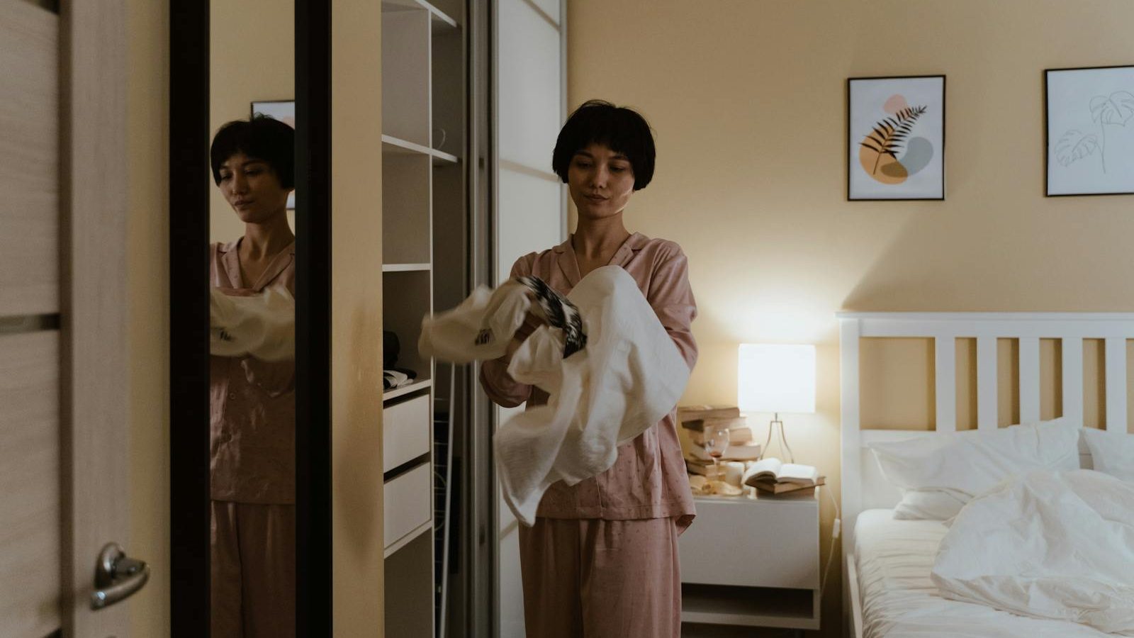 A woman in pajamas tidying up her closet in a warmly lit bedroom at night.