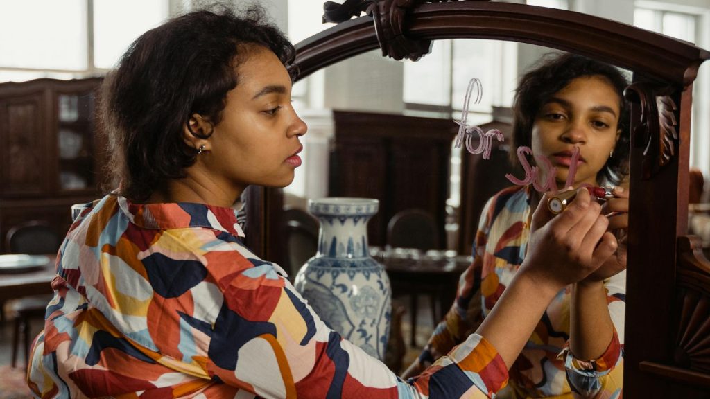 A woman writes 'for sale' on a mirror in an antique store, showcasing her reflection and vibrant printed shirt.