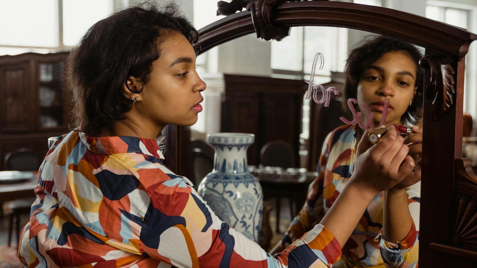 A woman writes 'for sale' on a mirror in an antique store, showcasing her reflection and vibrant printed shirt.