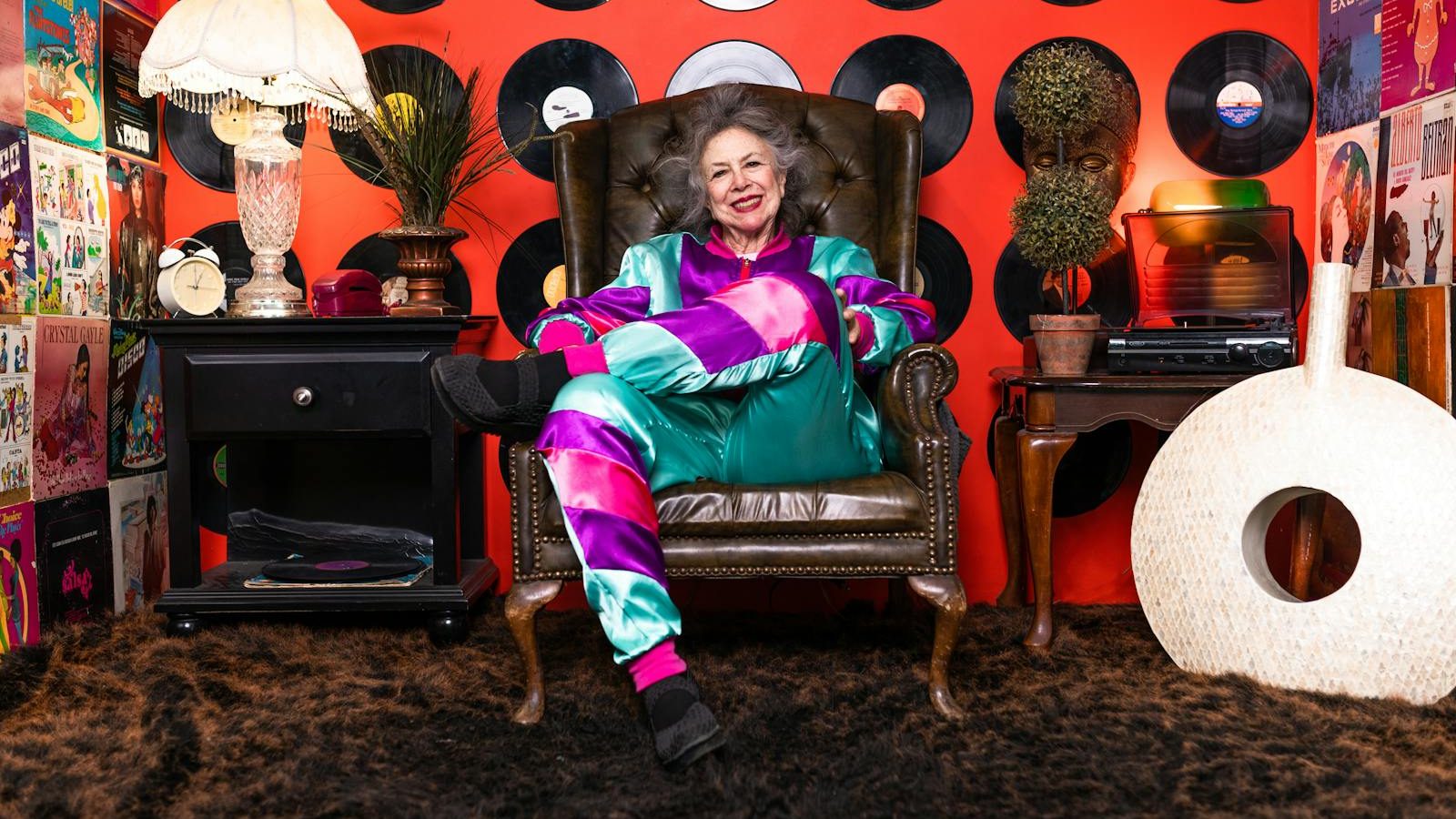 Elderly woman in colorful outfit relaxes in retro-themed room with vinyl records and vintage decor.