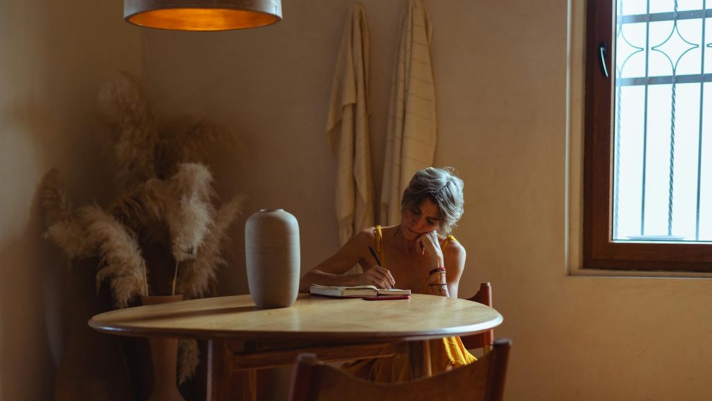 A senior woman enjoys journaling at a table in a warm, cozy home setting.