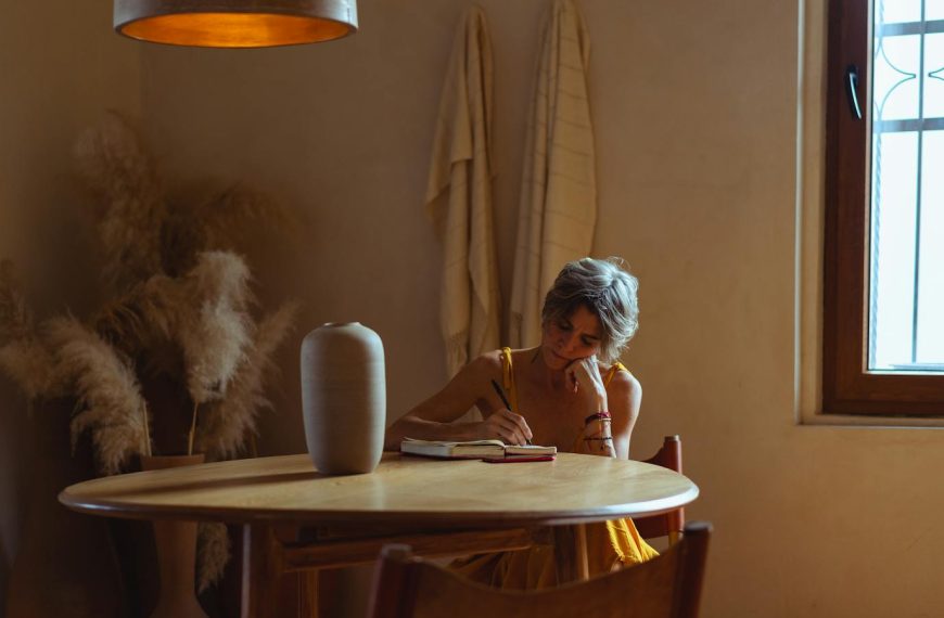 A senior woman enjoys journaling at a table in a warm, cozy home setting.