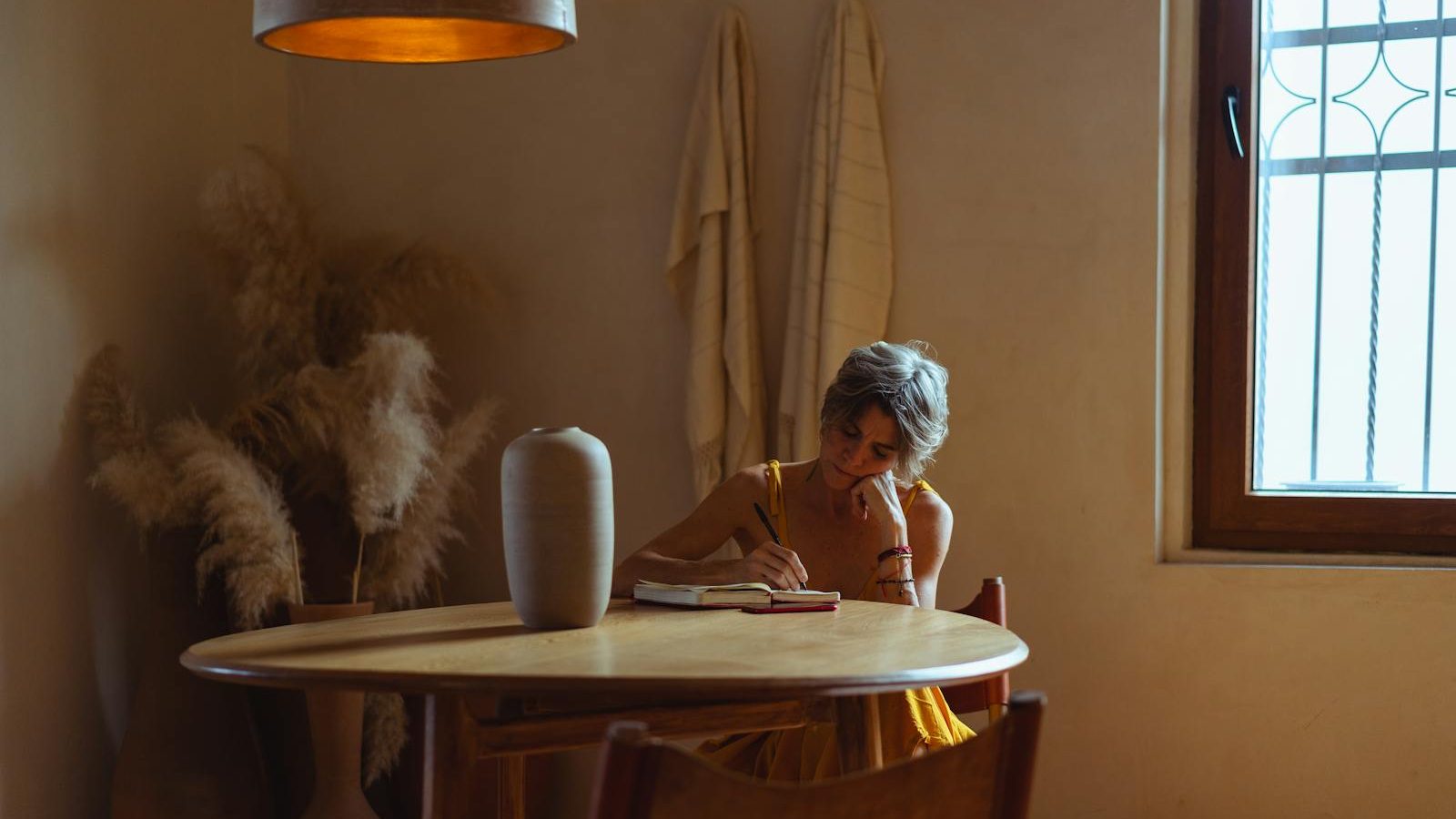 A senior woman enjoys journaling at a table in a warm, cozy home setting.