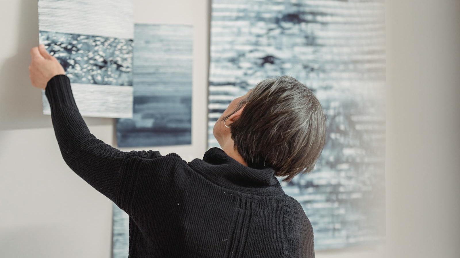 A woman arranges abstract art pieces on a gallery wall, showcasing modern creativity.