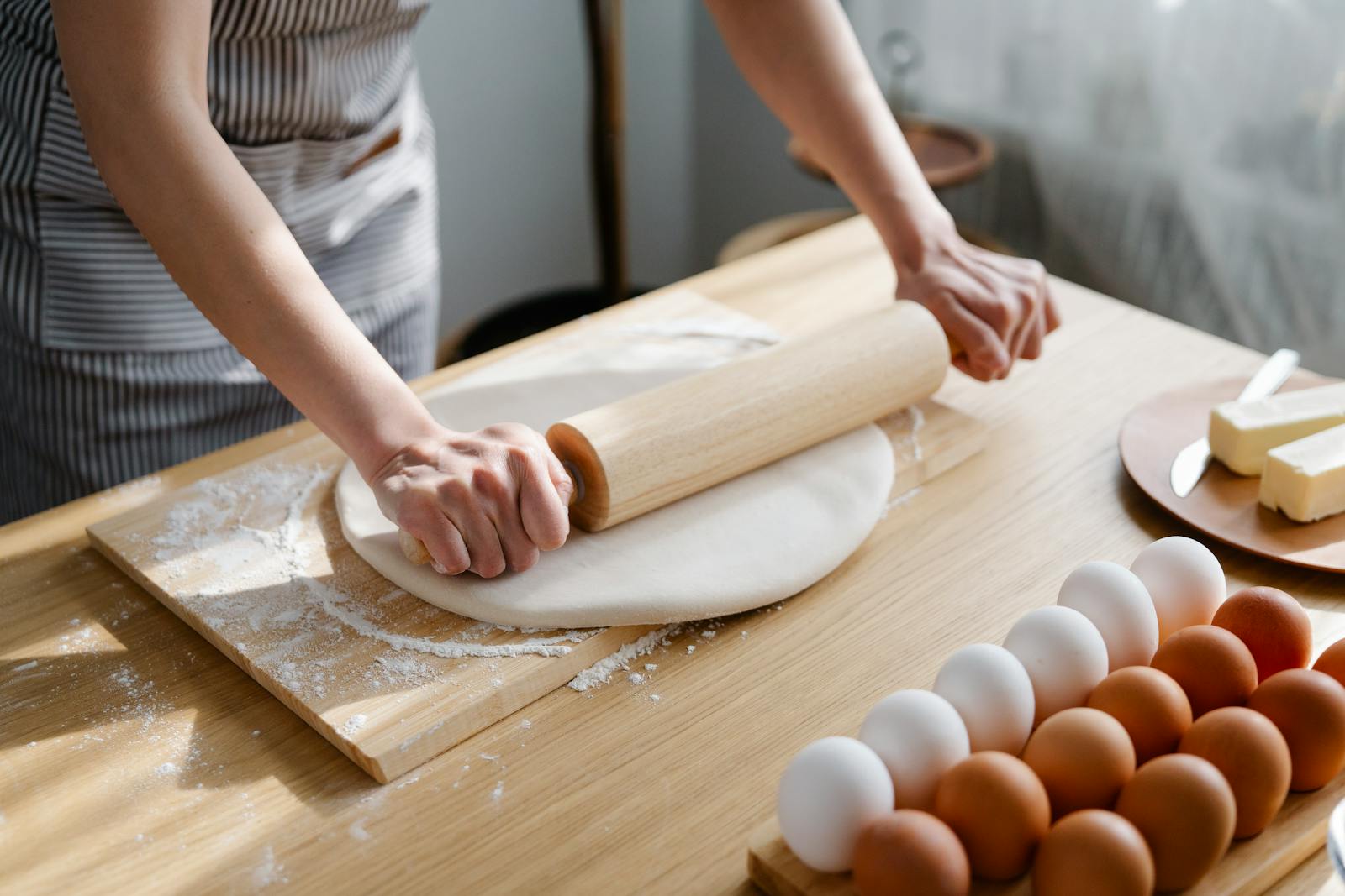A woman rolls out dough on a floured surface with eggs and butter nearby.