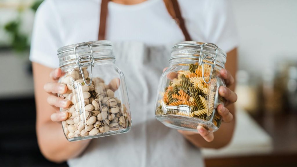 Hands holding glass jars filled with pistachios and colorful spiral pasta indoors.