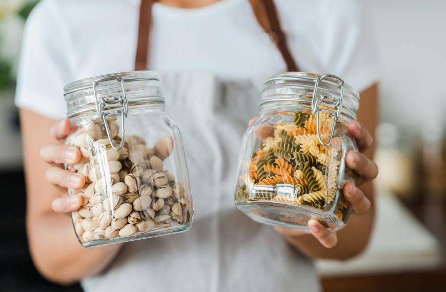 Hands holding glass jars filled with pistachios and colorful spiral pasta indoors.