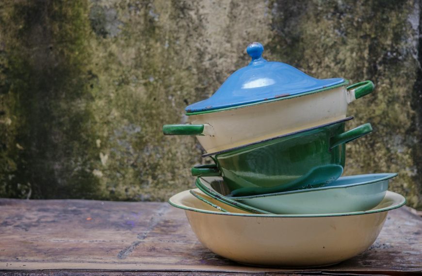 A rustic stack of colorful vintage enamel pots and pans outdoors against a textured wall.