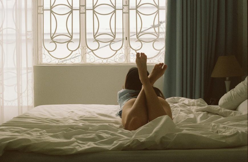 A woman relaxes on a bed with sunlight streaming through a modern bedroom window.