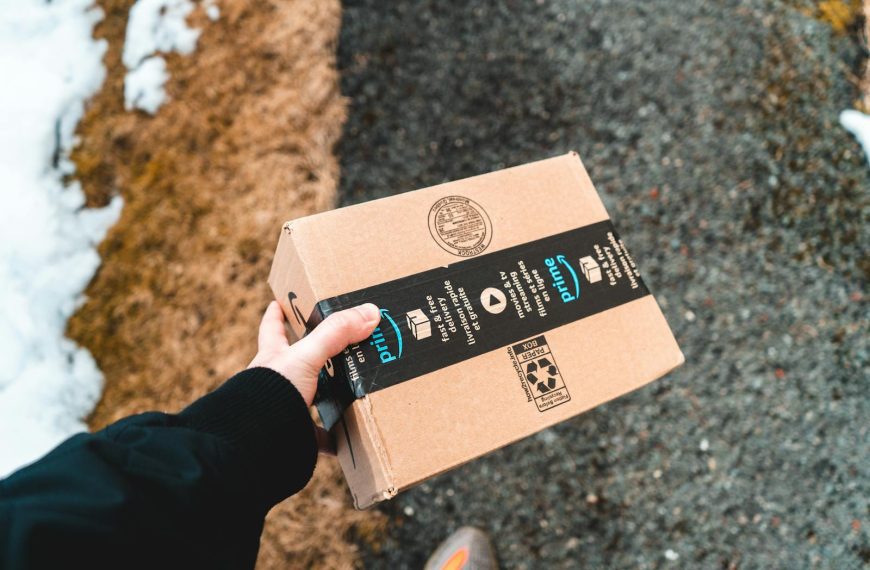 A person holding a cardboard Amazon Prime package on a snowy urban sidewalk.