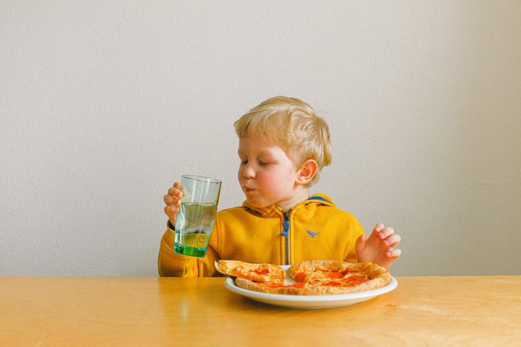 A cute blond child happily drinks while enjoying a pizza meal indoors.