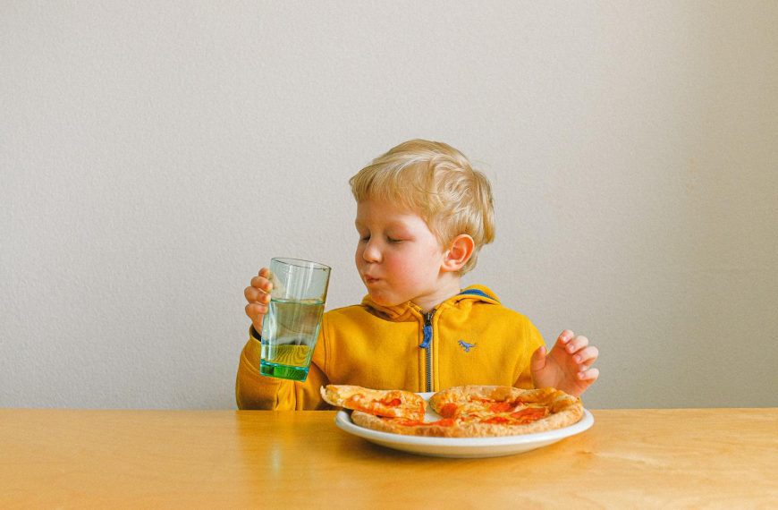 A cute blond child happily drinks while enjoying a pizza meal indoors.