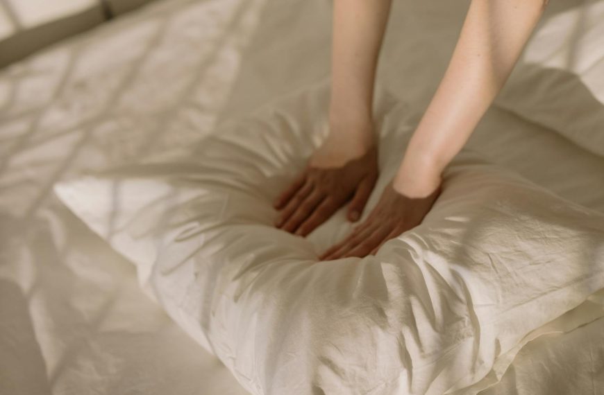 A warm, sunlit bedroom scene with hands fluffing a soft pillow, creating a cozy atmosphere.