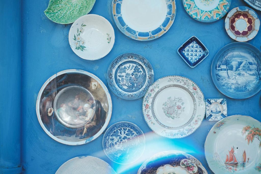 Assorted vintage plates of different sizes and ornaments hanging on bright blue wall in light studio