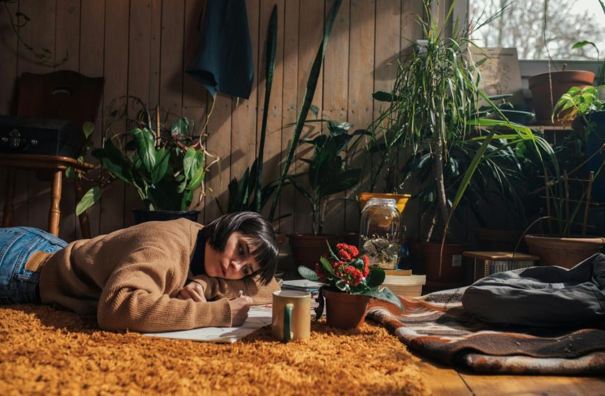 Adult woman drawing on a carpeted floor surrounded by indoor plants in cozy natural light.
