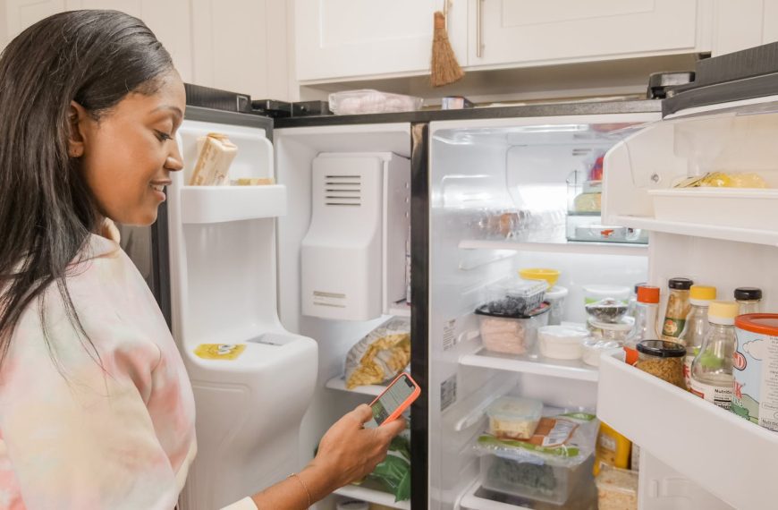 A woman stands at the open fridge in a kitchen, checking her phone.