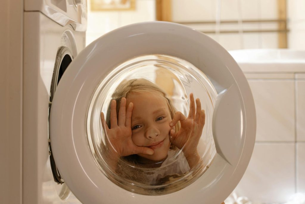 A child making funny faces behind a washing machine door in a playful indoor setting.