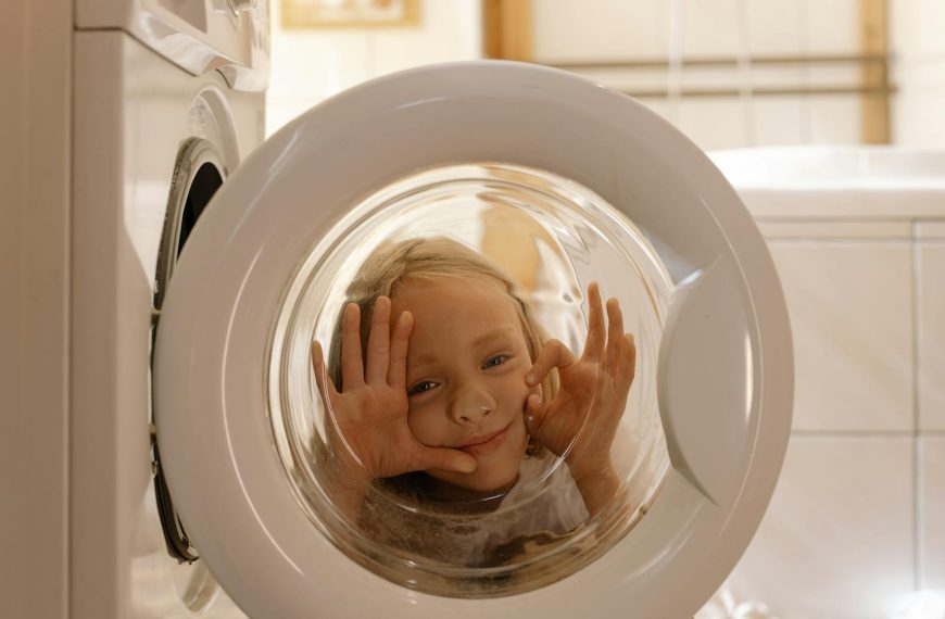 A child making funny faces behind a washing machine door in a playful indoor setting.
